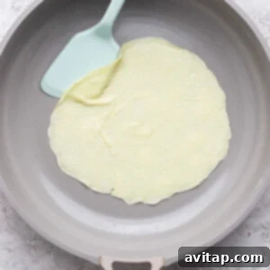 Creamy Cottage Cheese Crepes 10 A thin crepe cooking in a non-stick pan with a blue spatula lifting the edge.