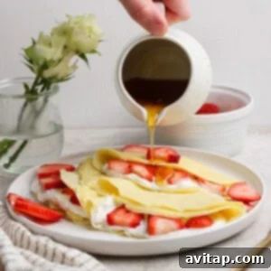 Creamy Cottage Cheese Crepes 11 A hand pours syrup over crepes filled with strawberries and cream, placed on a white plate. A small vase with white roses and a striped cloth are in the background.