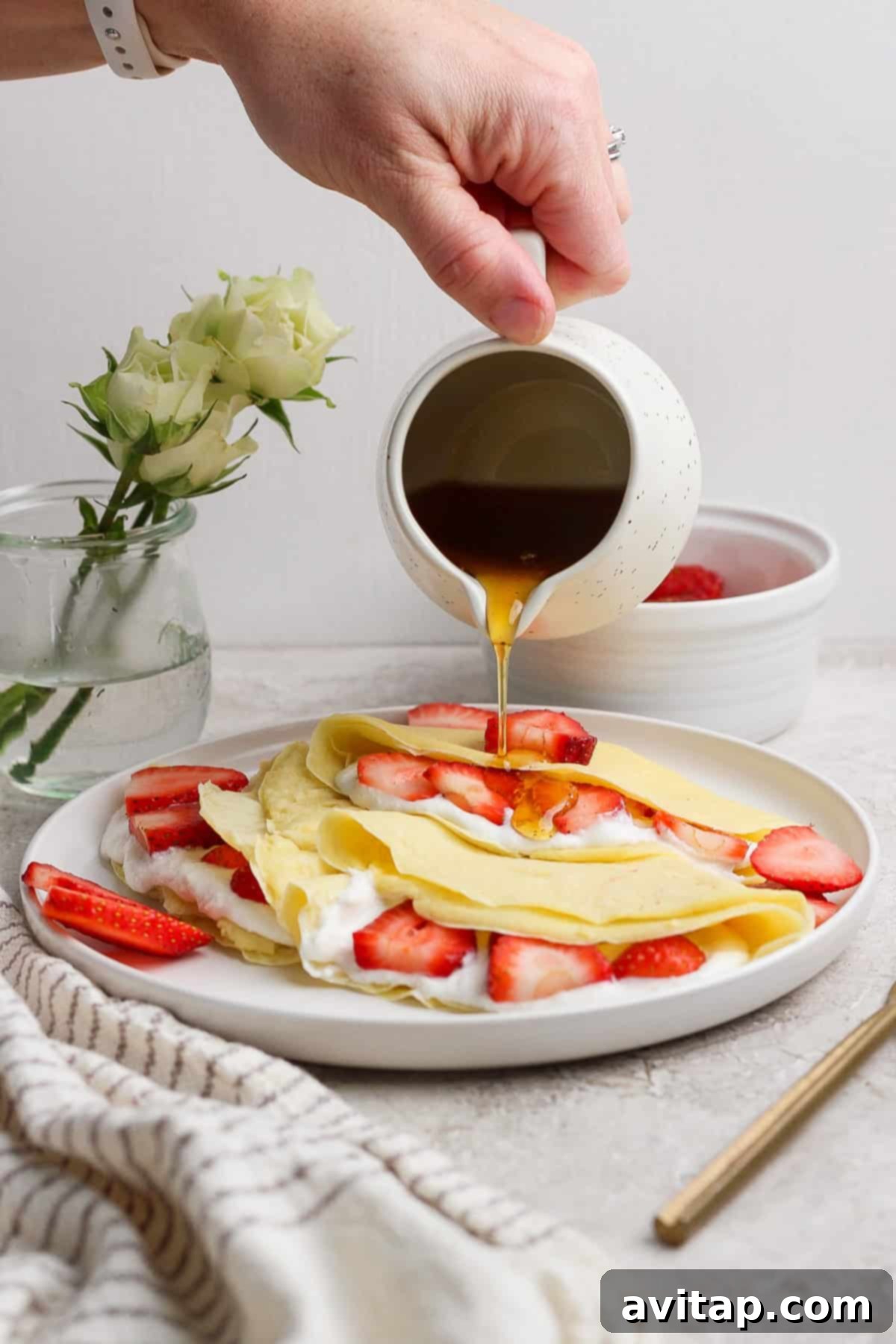 Creamy Cottage Cheese Crepes 5 A close-up shot of a hand gracefully pouring golden maple syrup over delicate crepes, generously filled with fresh strawberries and a creamy topping, all presented beautifully on a white plate. In the soft-focus background, a small vase of pristine white roses and a textured striped cloth add to the serene and inviting atmosphere.