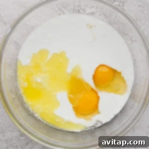 Creamy Cottage Cheese Crepes 8 A glass bowl containing two cracked eggs and milk, set on a textured gray surface.