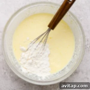 Creamy Cottage Cheese Crepes 9 A whisk in a glass bowl mixes flour into a yellow batter on a light textured surface.