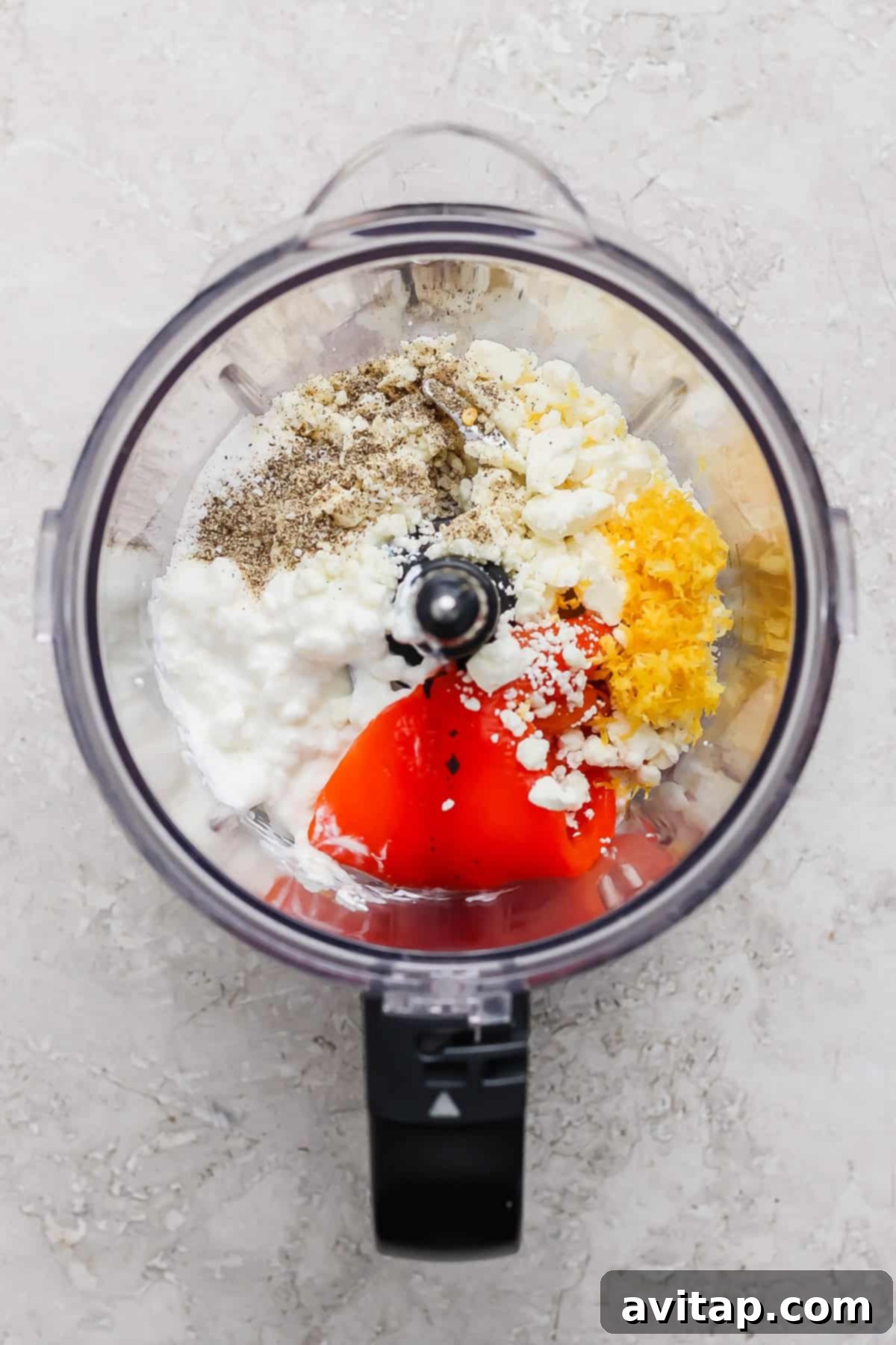 Ingredients including pepper, feta cheese, cottage cheese, lemon zest, and red peppers in a food processor, ready to be blended.