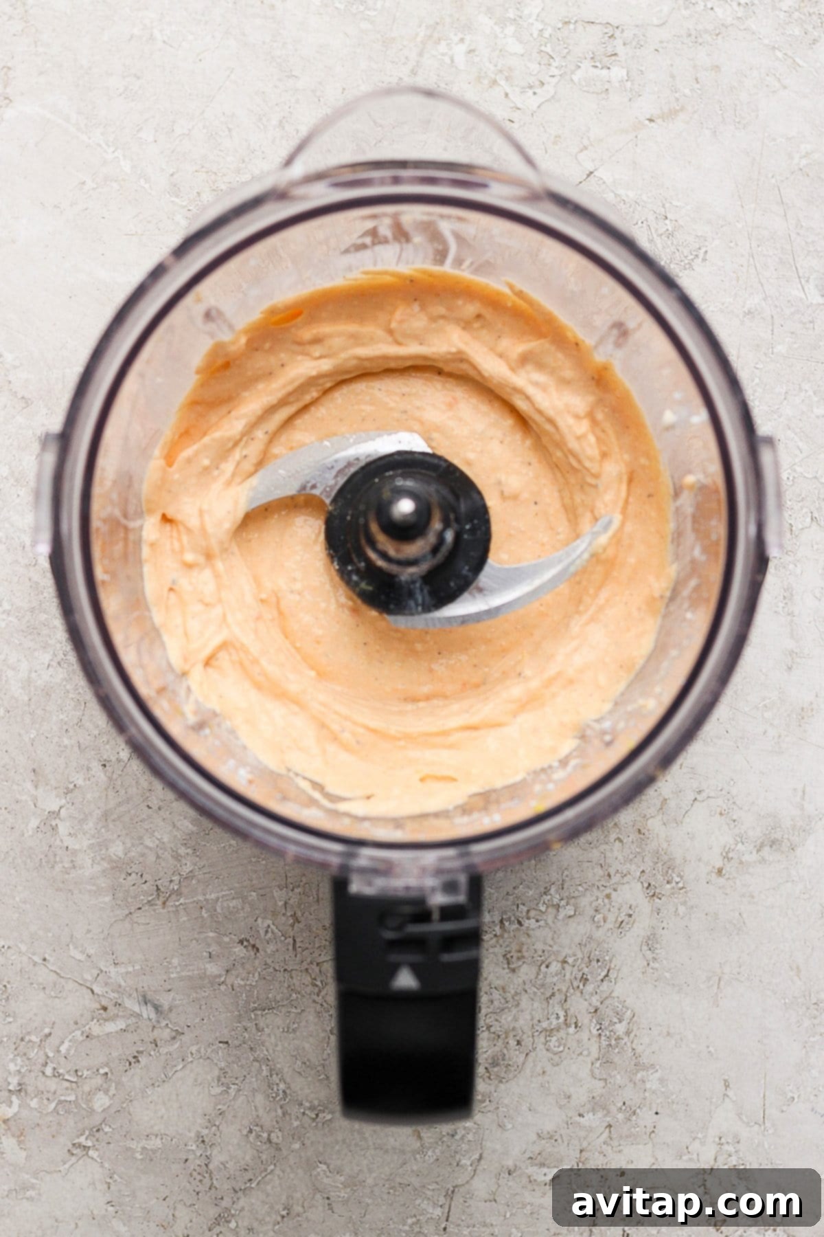 Food processor with creamy orange hummus on a gray textured surface.