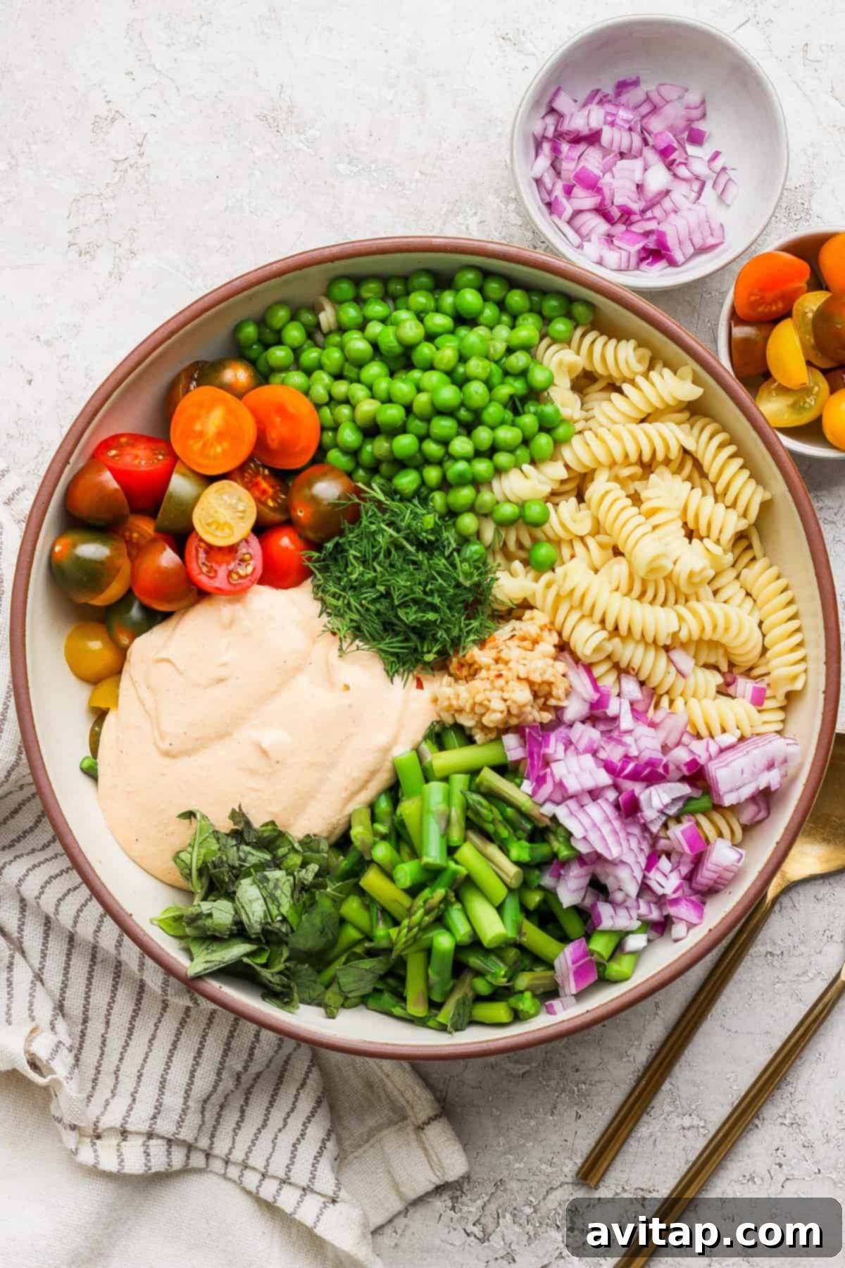 A bowl with rotini pasta, cherry tomatoes, peas, chopped red onion, green beans, fresh herbs, and a creamy sauce. A small bowl of onions and cutlery are beside it.