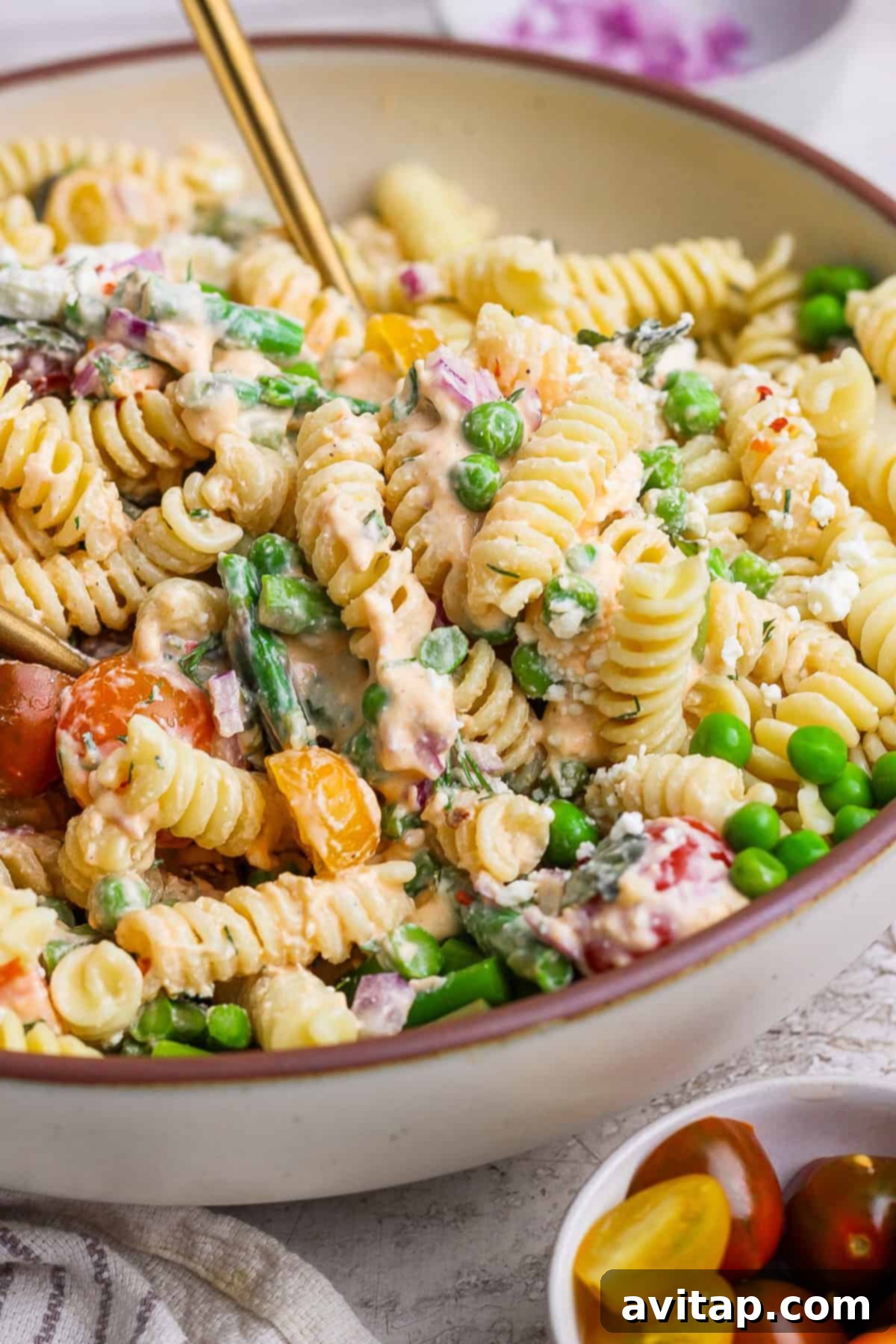 A bowl of rotini pasta salad with peas, asparagus, cherry tomatoes, and a creamy dressing.