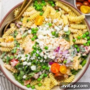 Bowl of pasta salad with rotini, peas, cherry tomatoes, crumbled cheese, greens, and a creamy dressing, garnished with chopped onions and red pepper flakes.