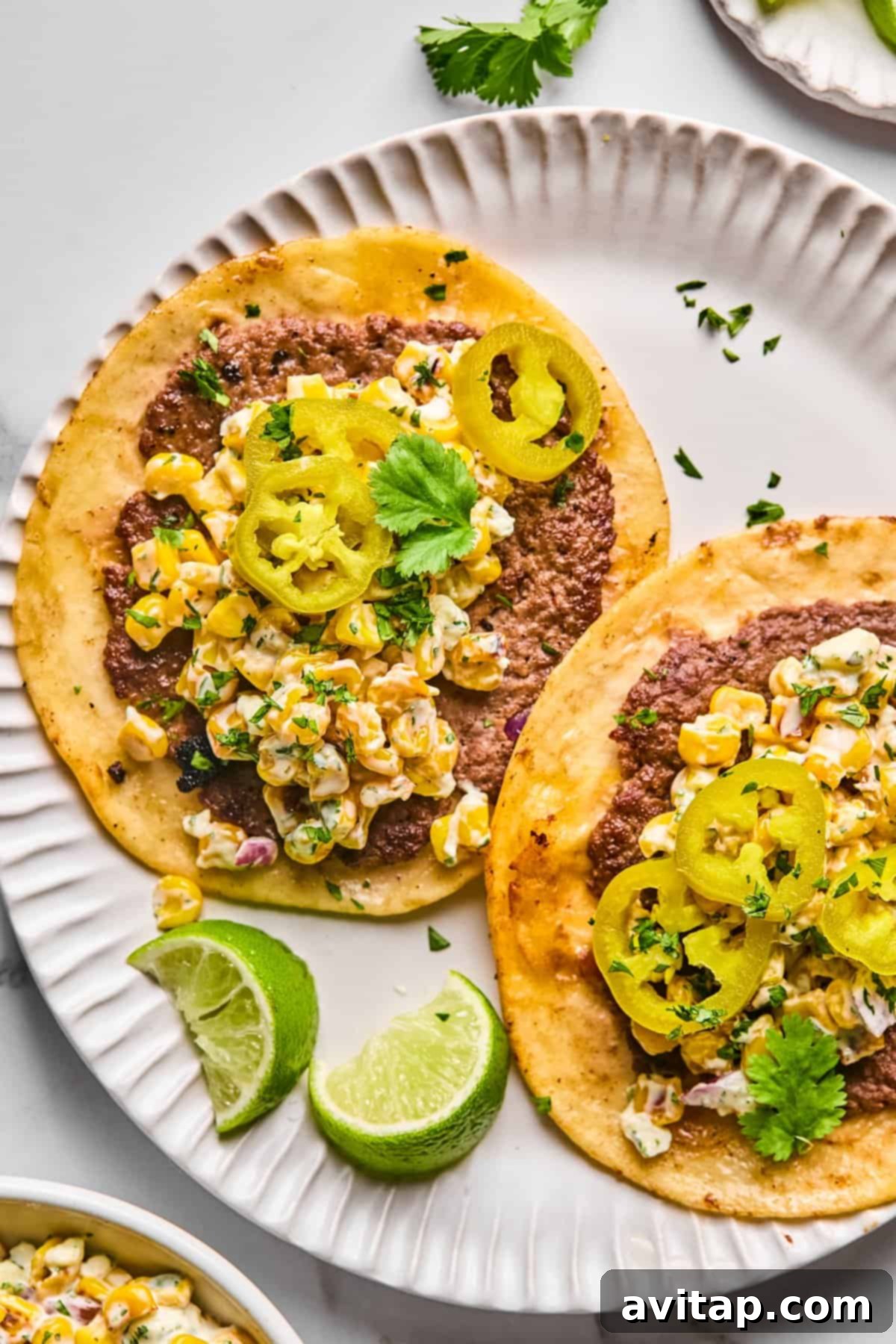 Two vibrant Street Corn Smash Tacos on a white plate, generously topped with seasoned ground beef, a creamy corn salad, bright green cilantro, and zesty pickled jalapeños, served with fresh lime wedges.
