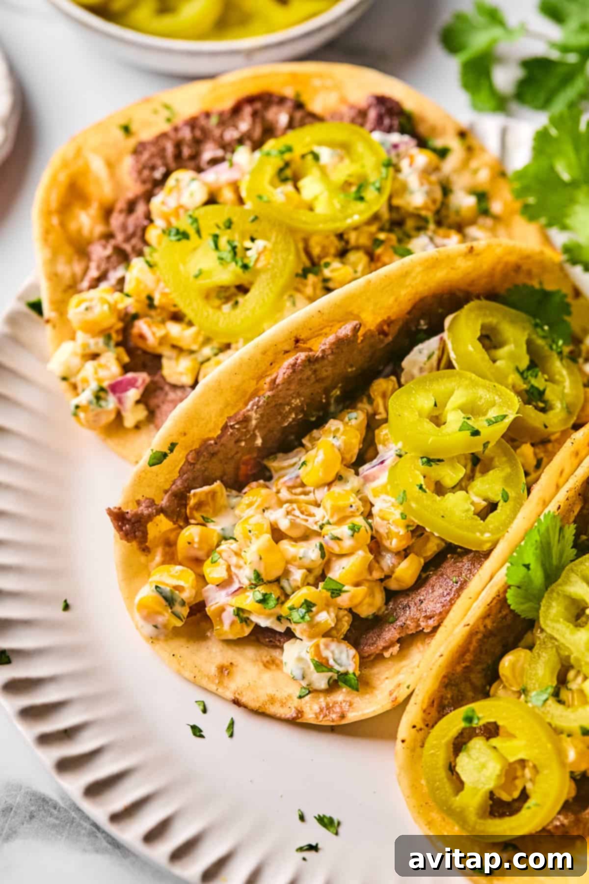 Two Street Corn Smash Tacos on a white plate, piled high with a flavorful corn salad, creamy refried beans, and garnished with vibrant pickled jalapeños and fresh herbs.
