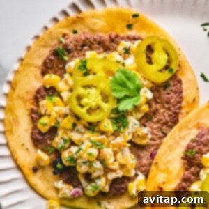 A close-up of a taco with ground beef, creamy corn salad, pickled jalapeño slices, and a sprig of cilantro on a white plate.
