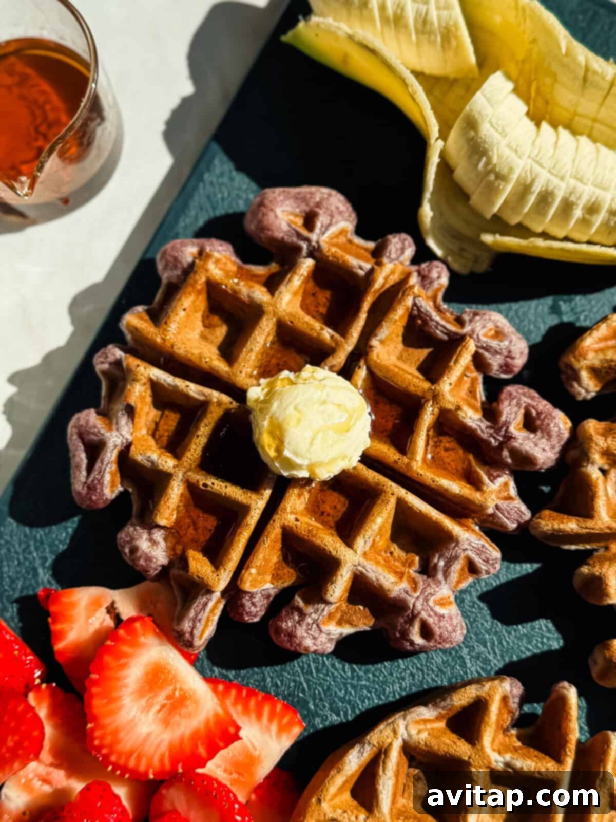A waffle topped with a pat of butter is shown alongside sliced bananas, strawberries, and a small glass of syrup on a dark surface.