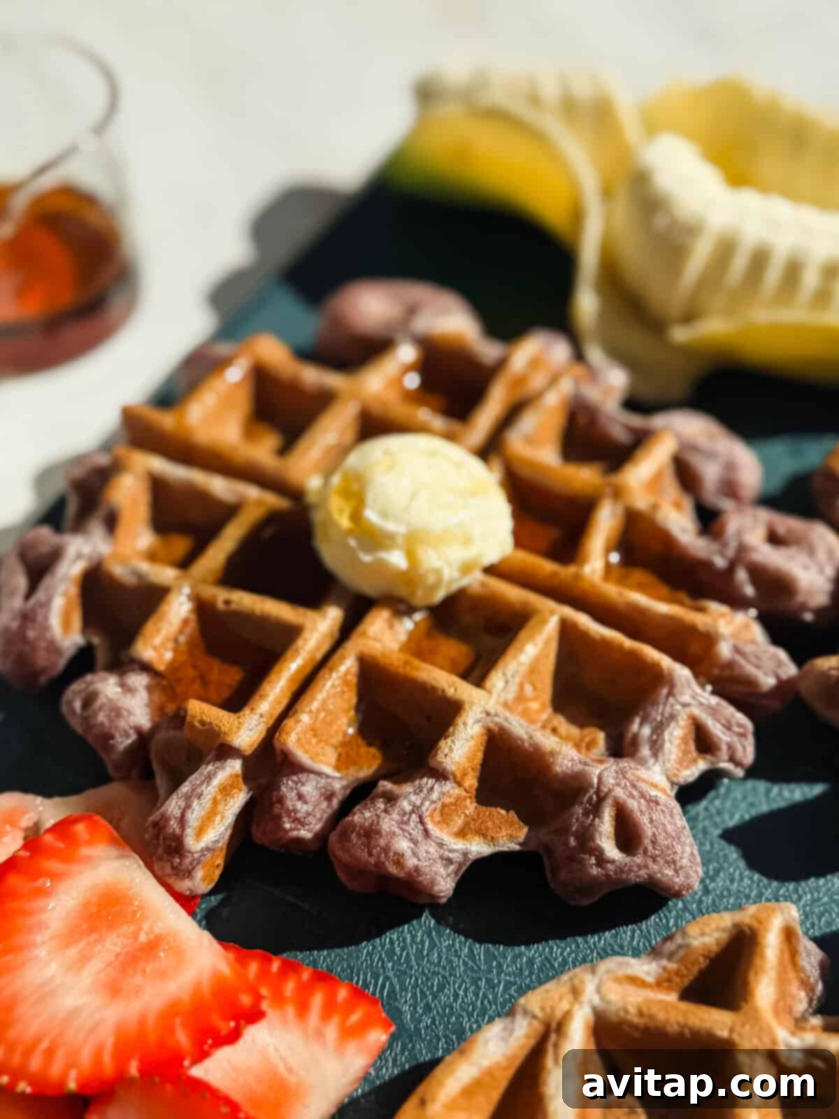 A close-up of a waffle topped with a scoop of butter and syrup, with sliced strawberries and a partially peeled banana in the background.