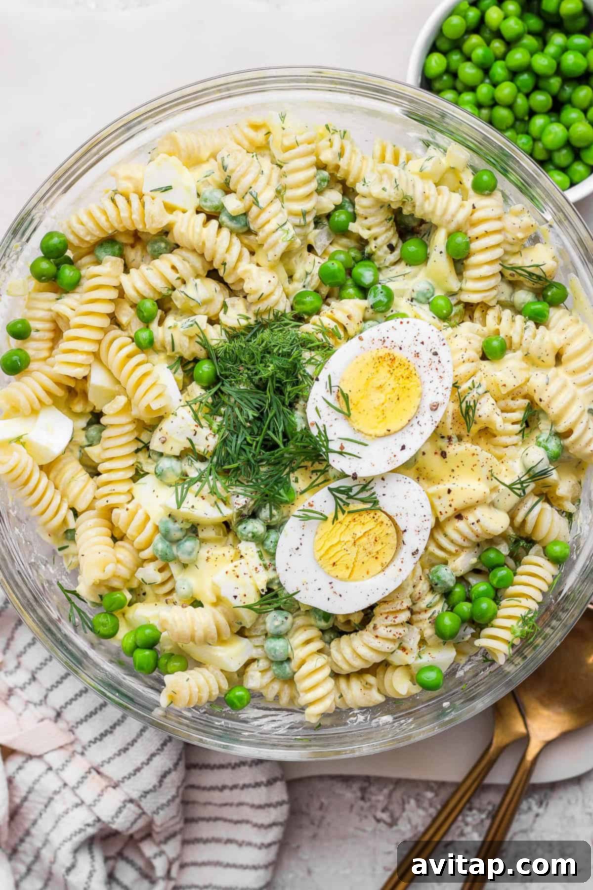 A vibrant bowl of deviled egg pasta salad featuring rotini pasta, green peas, sliced hard-boiled eggs, fresh dill, and a creamy, golden dressing. A gold spoon rests beside the bowl on a linen napkin, highlighting its appetizing appeal.