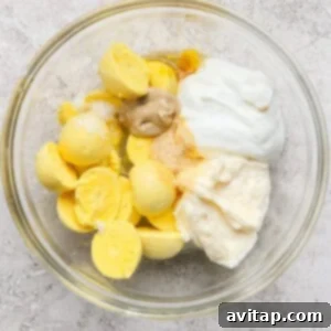 Bowl containing boiled egg yolks, mayonnaise, mustard, yogurt, and seasoning on a textured surface, ready to be mixed into a creamy dressing.