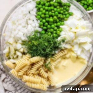 Glass bowl containing rotini pasta, peas, chopped onions, boiled eggs, pickles, fresh dill, and mayonnaise, set on a white surface with a silver spoon nearby, capturing a moment before mixing.