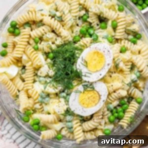 A bowl of pasta salad with rotini, green peas, hard-boiled egg slices, dill, and a creamy dressing. A gold spoon and a cloth napkin are next to the bowl, ready for serving.