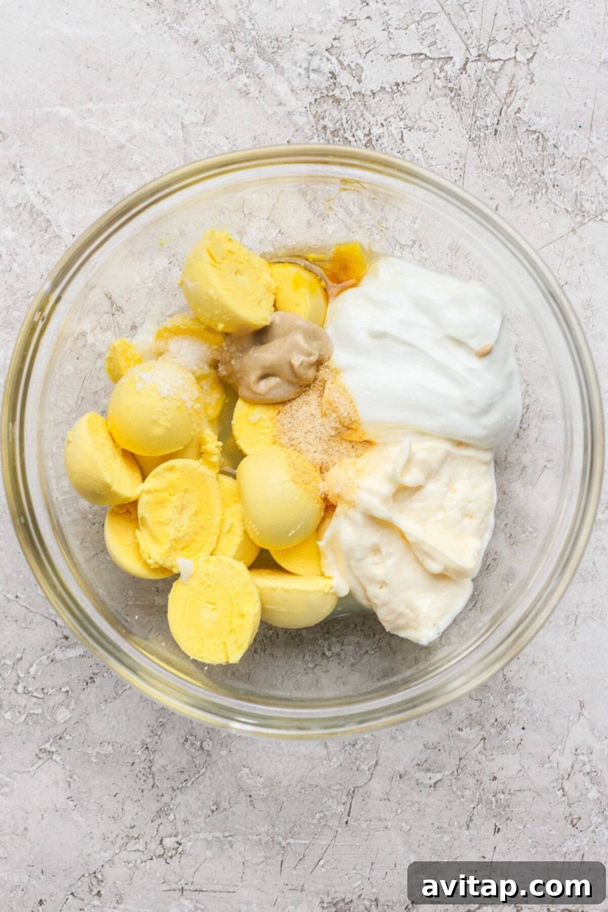 A glass bowl filled with mashed boiled egg yolks, blended with mayonnaise, mustard, Greek yogurt, and various seasonings, creating a creamy, golden mixture ready for the pasta salad.