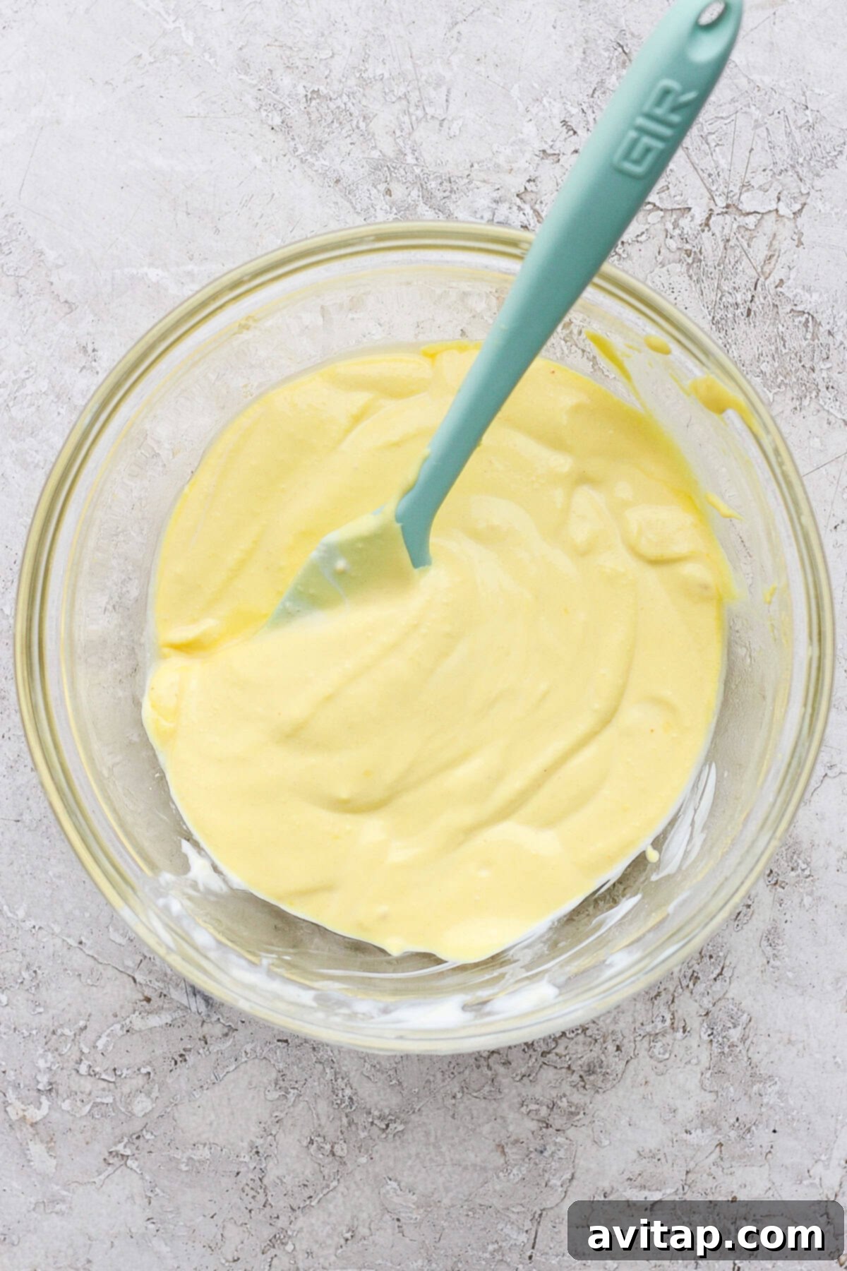 A teal spatula is seen stirring a smooth, creamy, and yellow mixture in a glass bowl set on a textured gray surface, showcasing the luscious consistency of the deviled egg sauce.