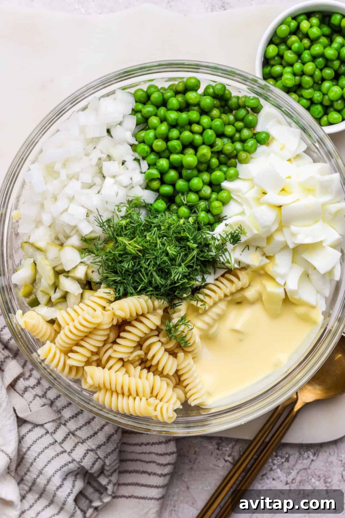 A spacious glass bowl is filled with a colorful array of ingredients for pasta salad, including rotini pasta, vibrant green peas, finely chopped onions, chunky pieces of boiled eggs, sliced dill pickles, and fresh dill sprigs, all waiting to be mixed with the creamy mayonnaise-based dressing. A silver spoon lies nearby on a pristine white surface.