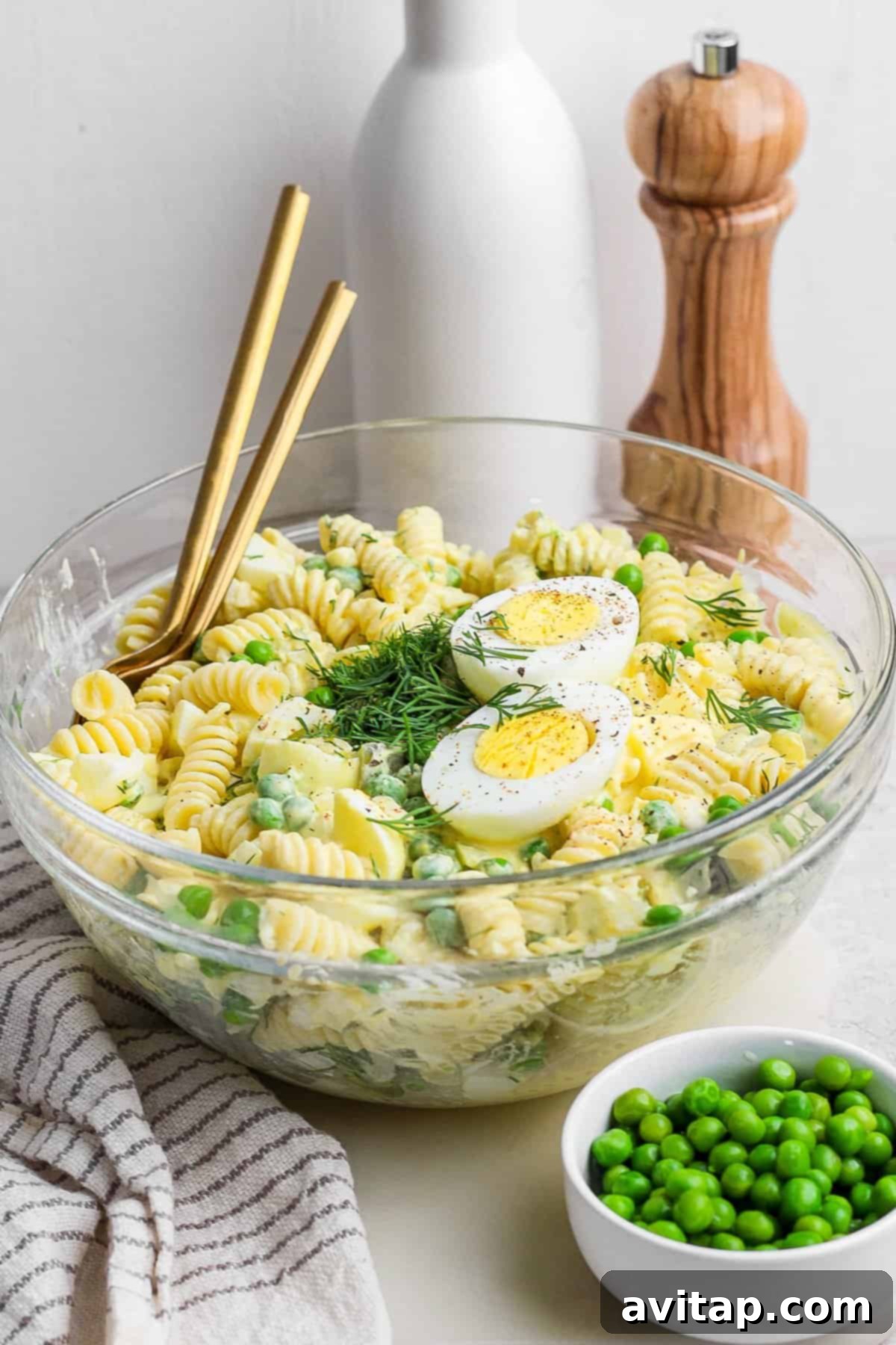 A beautifully arranged glass bowl of deviled egg pasta salad, featuring distinct slices of hard-boiled eggs and a generous amount of fresh herbs. It's elegantly placed on a table with a pair of chopsticks, a neatly folded white towel, a small bowl of fresh green peas, a sleek pepper mill, and an olive oil bottle, creating an inviting dining scene.
