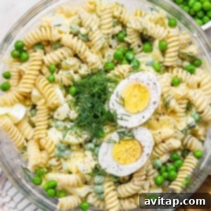 A bowl of fusilli pasta salad with green peas, chopped dill, and sliced hard-boiled egg halves on top.