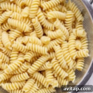 Cooked rotini pasta draining in a stainless steel colander over a sink, with droplets of water visible, signifying it's being rinsed with cold water.