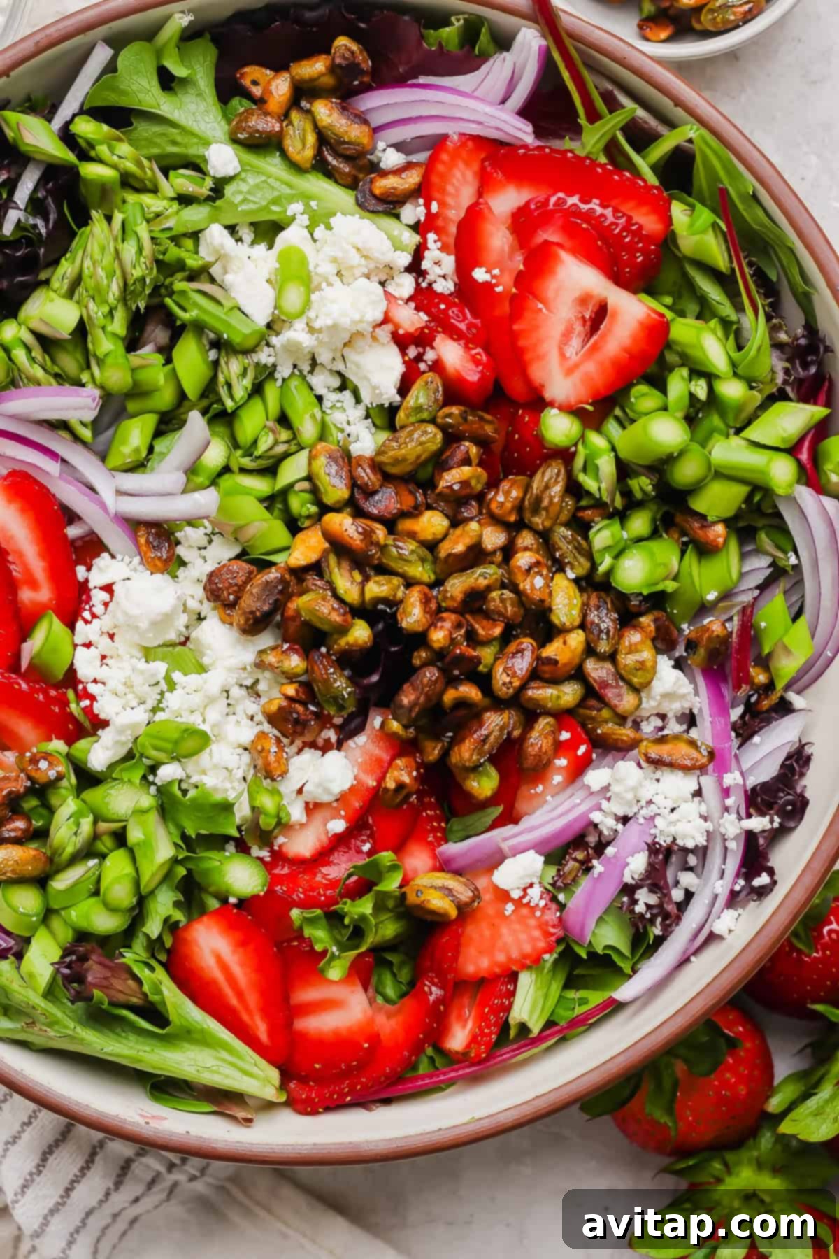 A vibrant summer salad featuring mixed greens, ruby-red sliced strawberries, thinly cut red onions, tender asparagus, green edamame, crumbled feta cheese, and glistening honey-roasted pistachios, all arranged beautifully in a large, inviting bowl.
