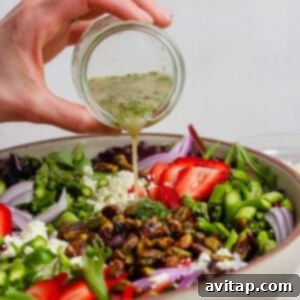 A hand pours vinaigrette from a small jar onto a vibrant salad with strawberries, feta cheese, red onions, pistachios, and greens in a large bowl, ready for serving.