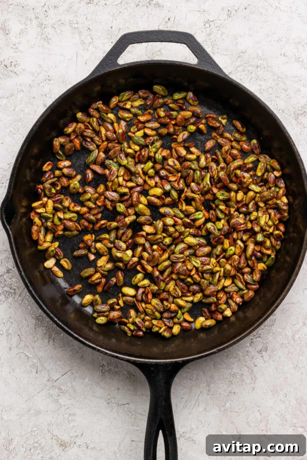A black cast iron skillet filled with glistening, golden-brown honey-roasted pistachios, resting on a light-textured kitchen surface.