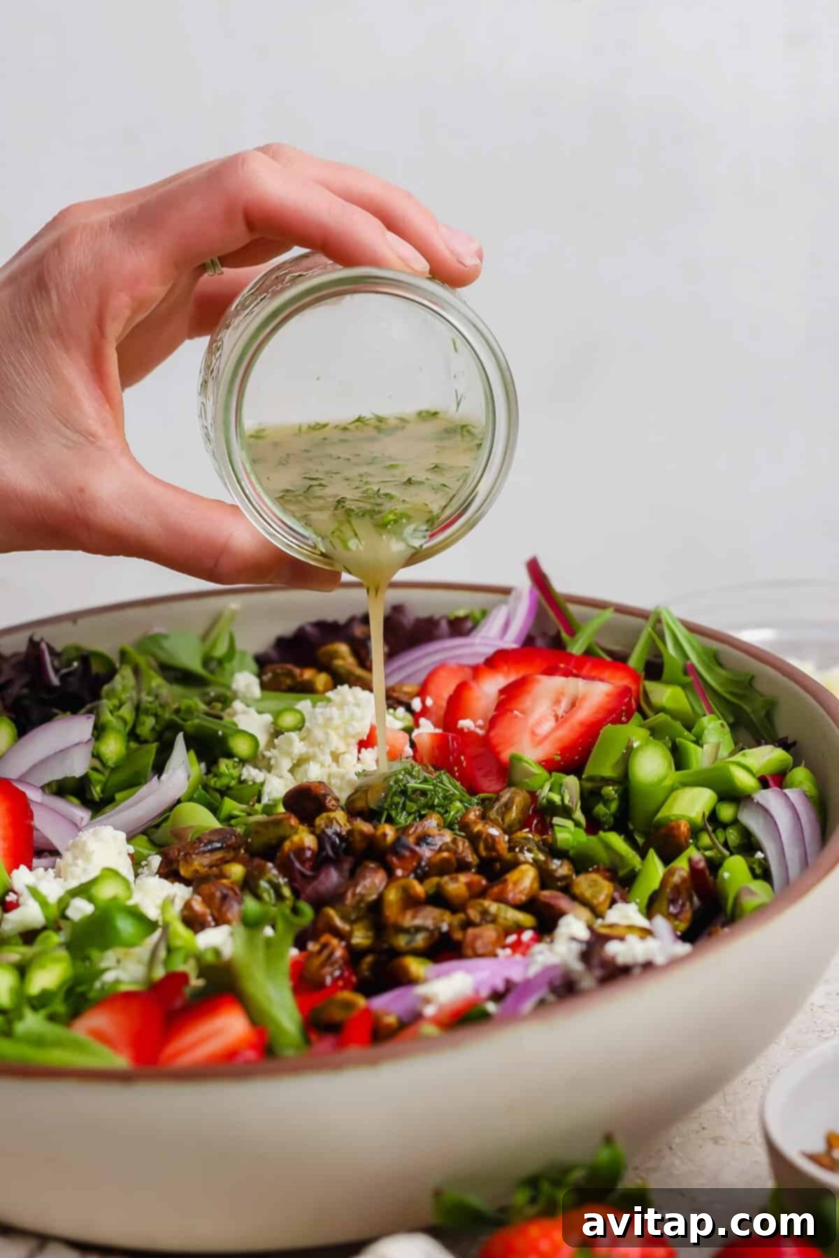 A hand gracefully pours a golden vinaigrette from a small glass jar onto a large, inviting bowl filled with a colorful salad. The salad features fresh strawberries, tangy feta cheese, thinly sliced red onions, crunchy pistachios, and a medley of vibrant greens, ready to be tossed.