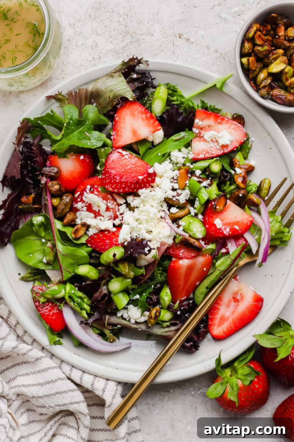 A beautifully plated summer salad featuring mixed greens, sweet sliced strawberries, savory crumbled feta cheese, crunchy honey-roasted pistachios, and thinly sliced red onion. A fork rests beside the plate, with a small bowl of raw pistachios and fresh strawberries in the background, hinting at the fresh ingredients.