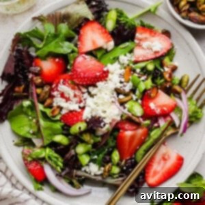 A plate of mixed green salad topped with sliced strawberries, crumbled feta cheese, red onion, pistachios, asparagus, and a gold fork on the side, showcasing the vibrant colors and textures of the dish.