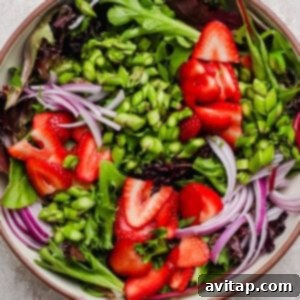 A bowl of mixed greens salad topped with sliced strawberries, red onion, and chopped green vegetables on a light textured surface, awaiting further additions.