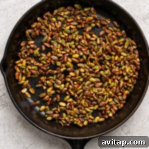 A black cast iron skillet filled with roasted pistachios, glistening with honey, sitting on a light textured surface as they caramelize.
