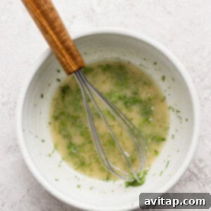A white bowl containing a light-colored vinaigrette with green herbs, being mixed with a metal whisk with a wooden handle, set on a light-textured surface, illustrating the dressing preparation.