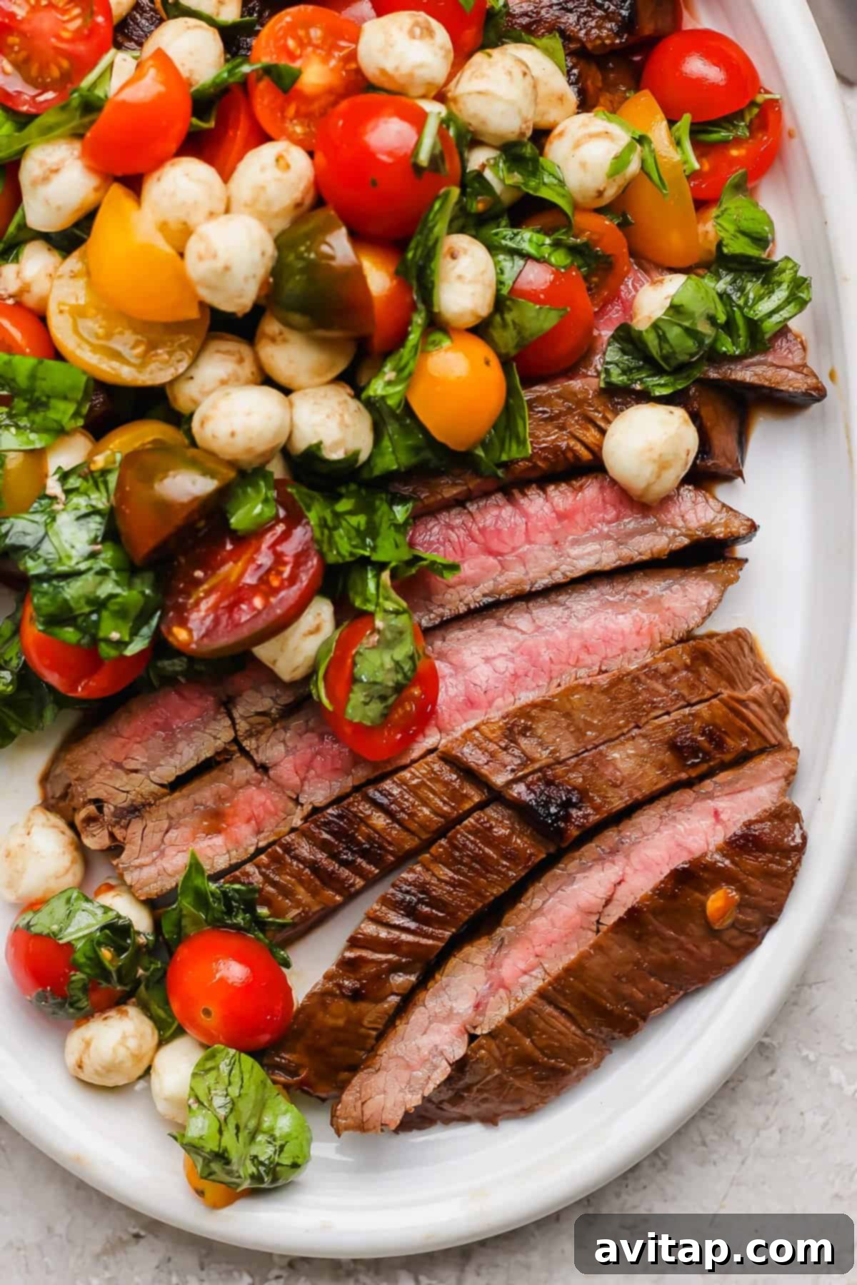 Sliced medium-rare balsamic skirt steak served with a vibrant caprese salad of cherry tomatoes, mozzarella balls, and fresh basil on a pristine white plate, showcasing a perfect summer meal.