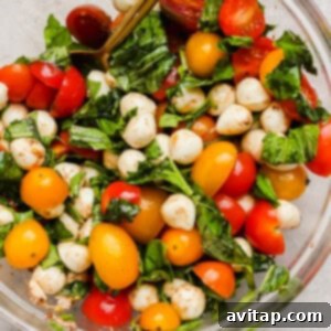 A clear glass bowl brimming with a colorful Caprese salad, featuring bright cherry tomatoes, creamy mozzarella balls, and aromatic fresh basil leaves, along with a spoon, poised on a light surface.