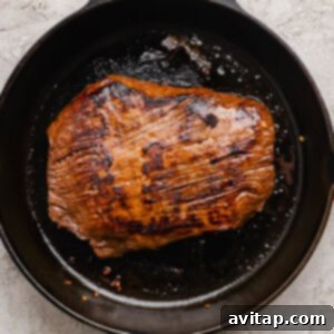 A large piece of marinated steak actively searing in a black cast iron skillet, producing a delicious crust on a light textured surface.