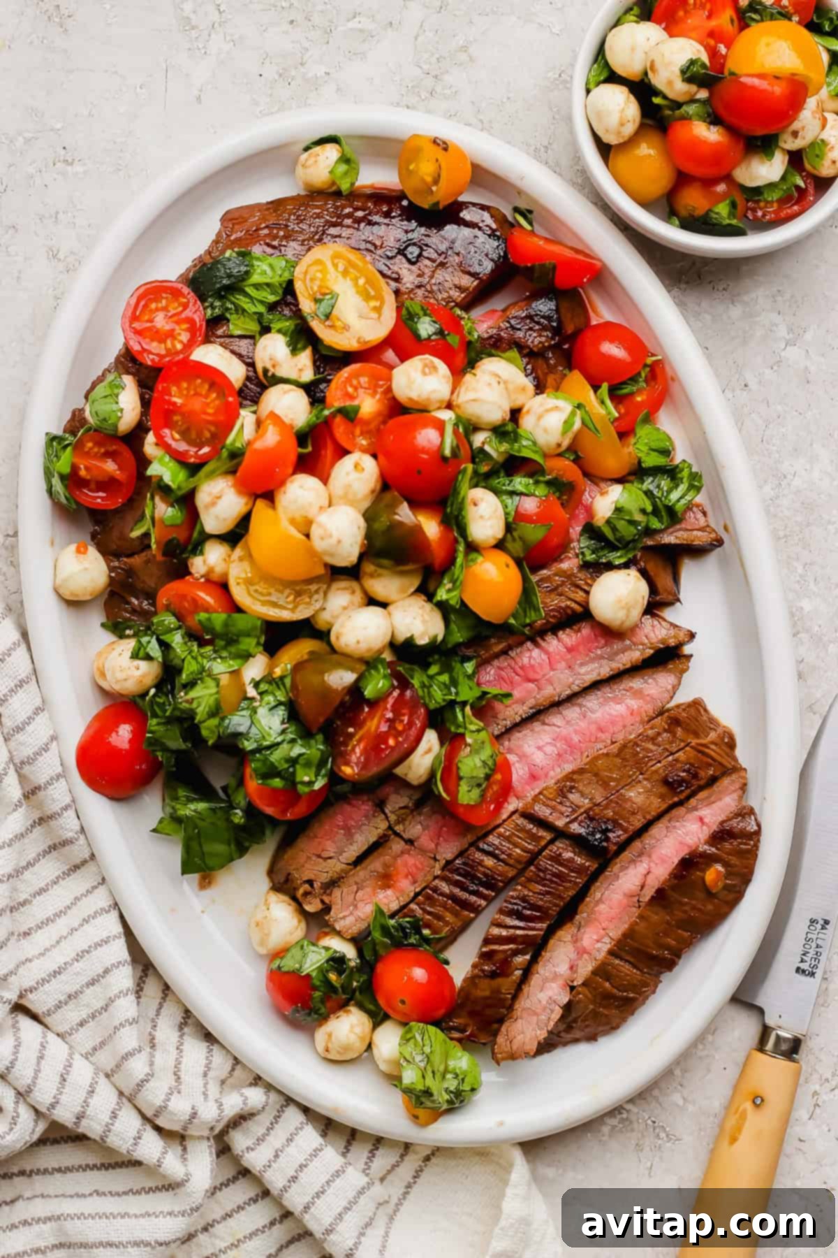 A beautifully arranged oval platter features tender, sliced medium-rare balsamic skirt steak, generously topped with a fresh Caprese salad of bright tomatoes, creamy mozzarella, and fragrant basil. A smaller matching bowl of caprese, a striped linen towel, and a sharp knife complete the inviting presentation.