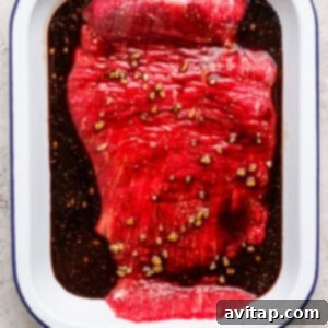 Raw flank steak marinating in a white dish with a rich, dark balsamic sauce and visible minced garlic, set on a light textured surface.