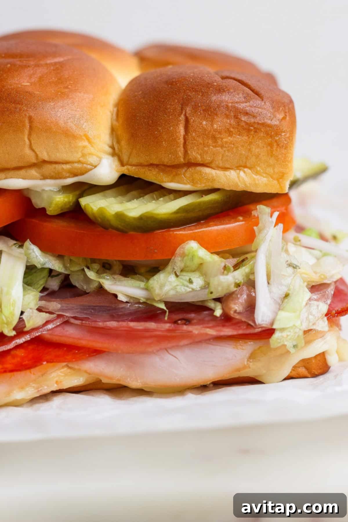 A close-up of a perfectly layered Italian Grinder Slider on a soft Hawaiian roll, showcasing sliced meats, shredded lettuce, ripe tomato, crisp pickles, and a creamy mayonnaise spread.