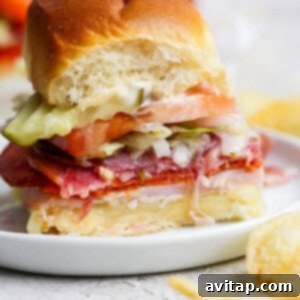 A small, perfectly assembled Italian Grinder Slider on a white plate, layered with deli meats, melted cheese, fresh lettuce, tomato, and pickles, with a bite taken out. Crispy potato chips are visible beside the plate, completing the meal.
