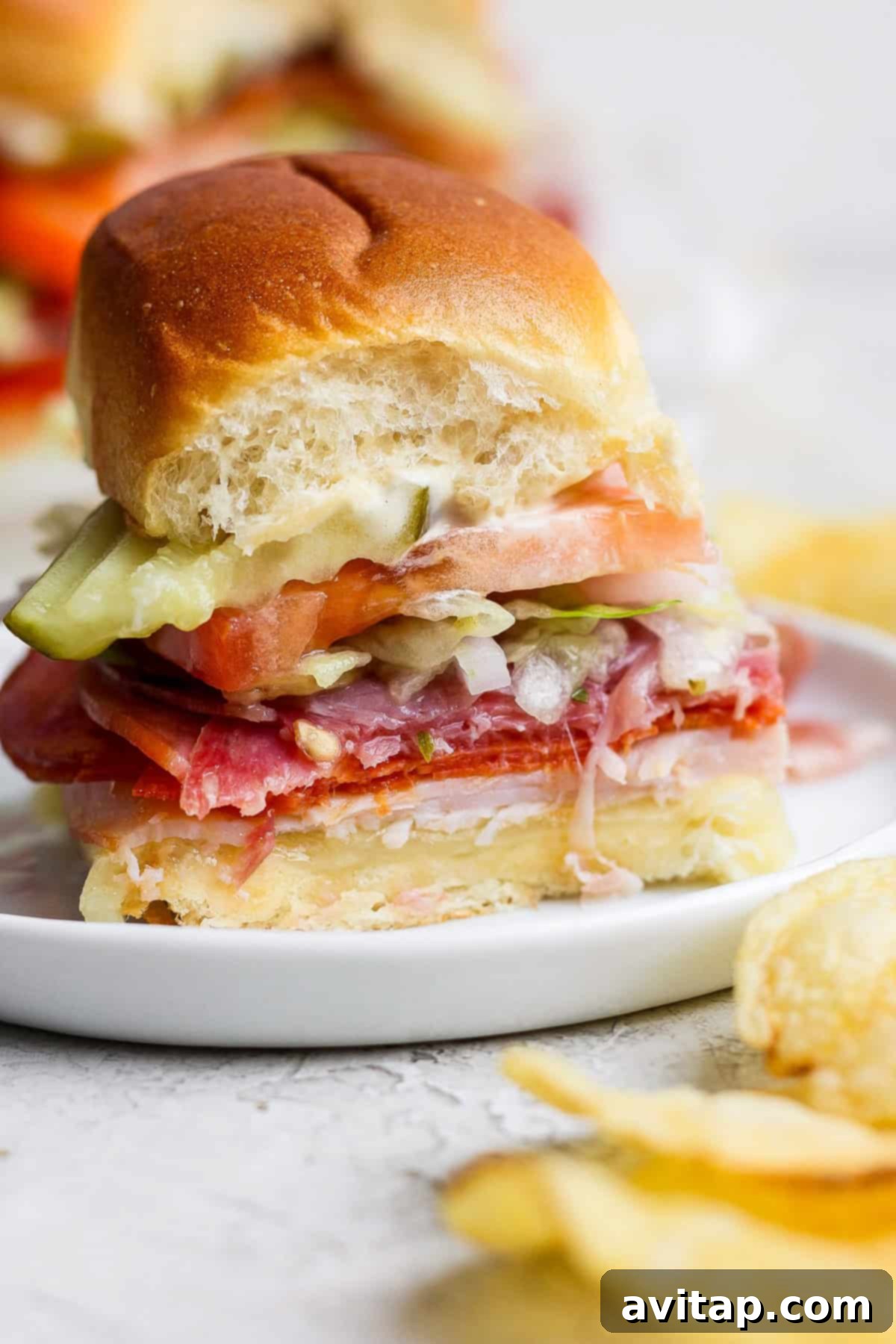 A freshly cut Italian Grinder Slider on a white plate, showcasing all the layers: deli meats, cheese, lettuce, tomato, and pickles. Potato chips are visible beside the plate, ready for serving.