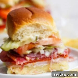 A small sandwich with sliced meats, cheese, tomato, lettuce, pickles, and mayonnaise on a soft bun, shown with a bite taken out of it.