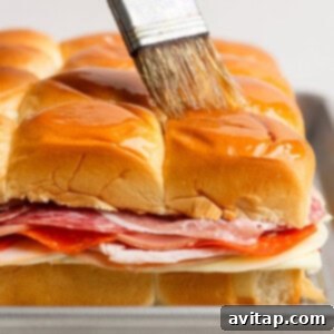 A hand brushes melted butter over the tops of stacked sandwich sliders with layers of deli meats and cheese on a baking tray, ready for the oven.