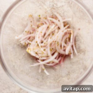 A clear glass bowl containing thinly sliced white onions mixed with red wine vinegar and dried oregano, marinating to create the zesty grinder salad base.