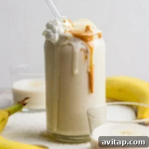 Glass of banana smoothie topped with whipped cream and peanut butter, surrounded by bananas and two small glasses of smoothie.
