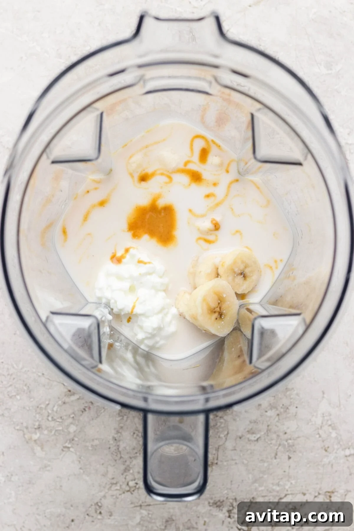 A blender containing milk, sliced banana, cottage cheese, and a drizzle of honey, viewed from above.