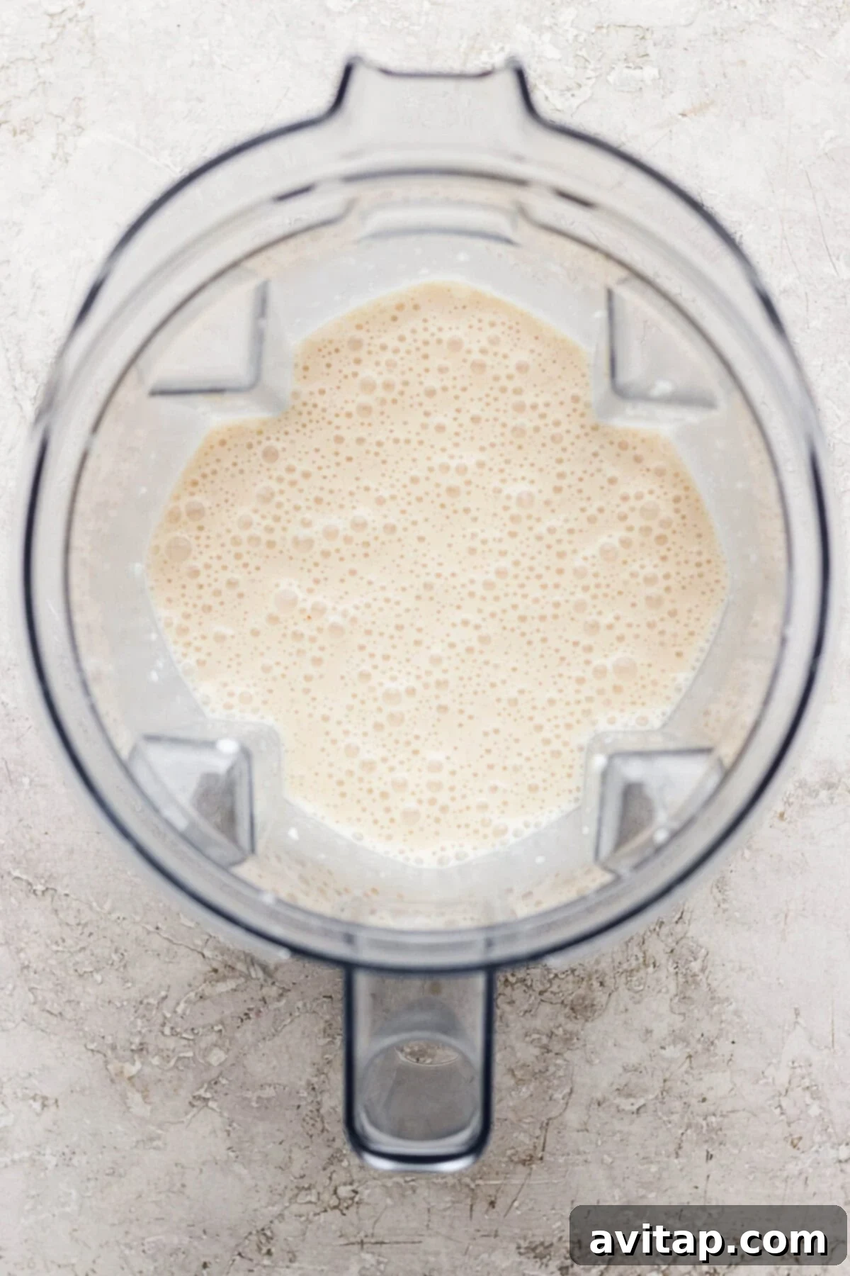 Top-down view of a blender containing a light beige, frothy liquid on a light textured surface.