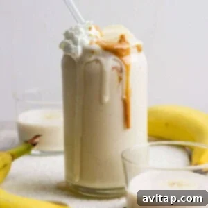 A glass of peanut butter banana protein shake topped with whipped cream, banana slices, and peanut butter, with a straw, surrounded by bananas and two smaller glasses of smoothie.