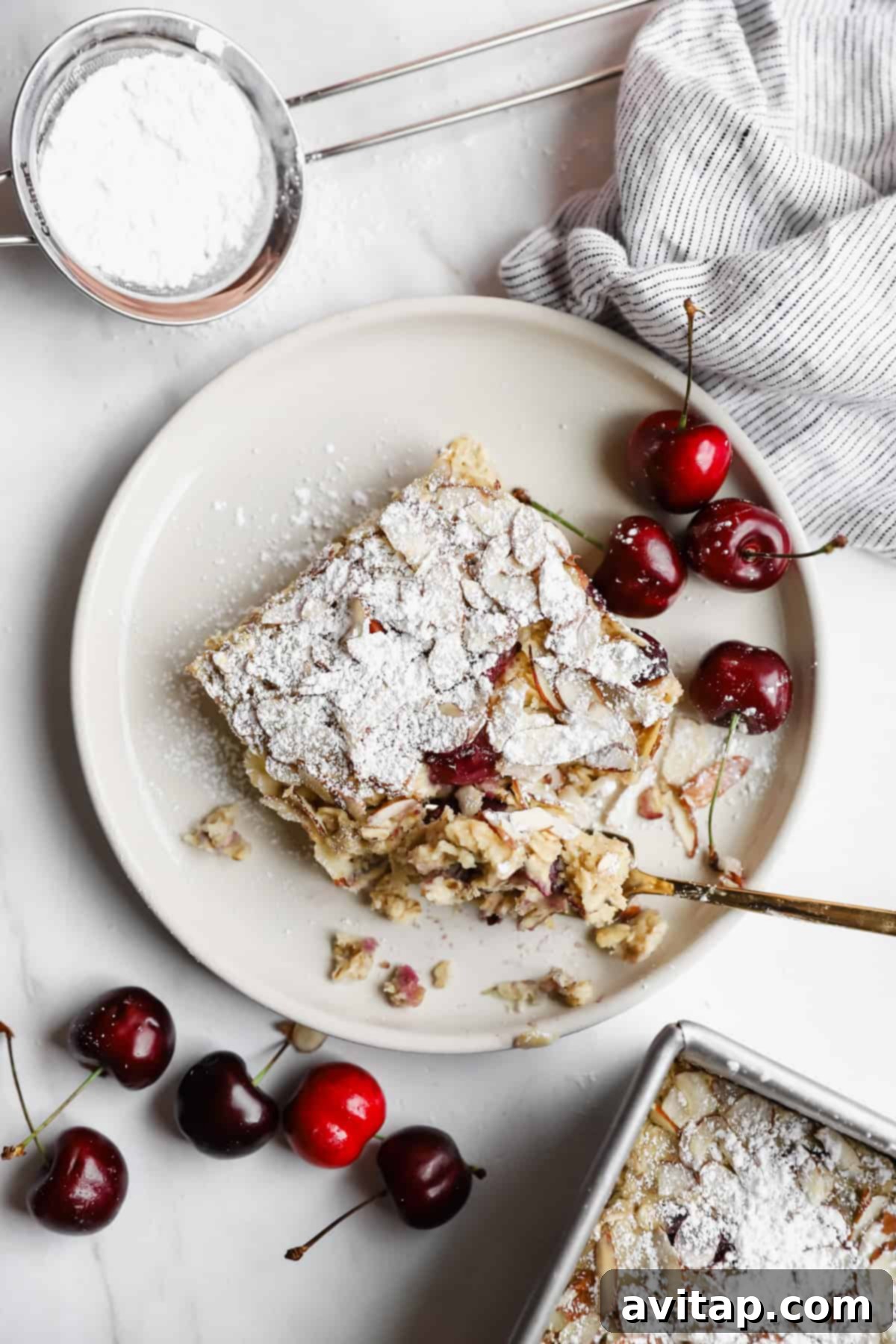 A perfectly baked slice of cherry almond croissant baked oats, lightly dusted with powdered sugar, resting on a white plate. Fresh, ruby-red cherries are scattered around the plate, alongside a striped linen cloth and a small sifter with powdered sugar, creating an inviting and picturesque scene.